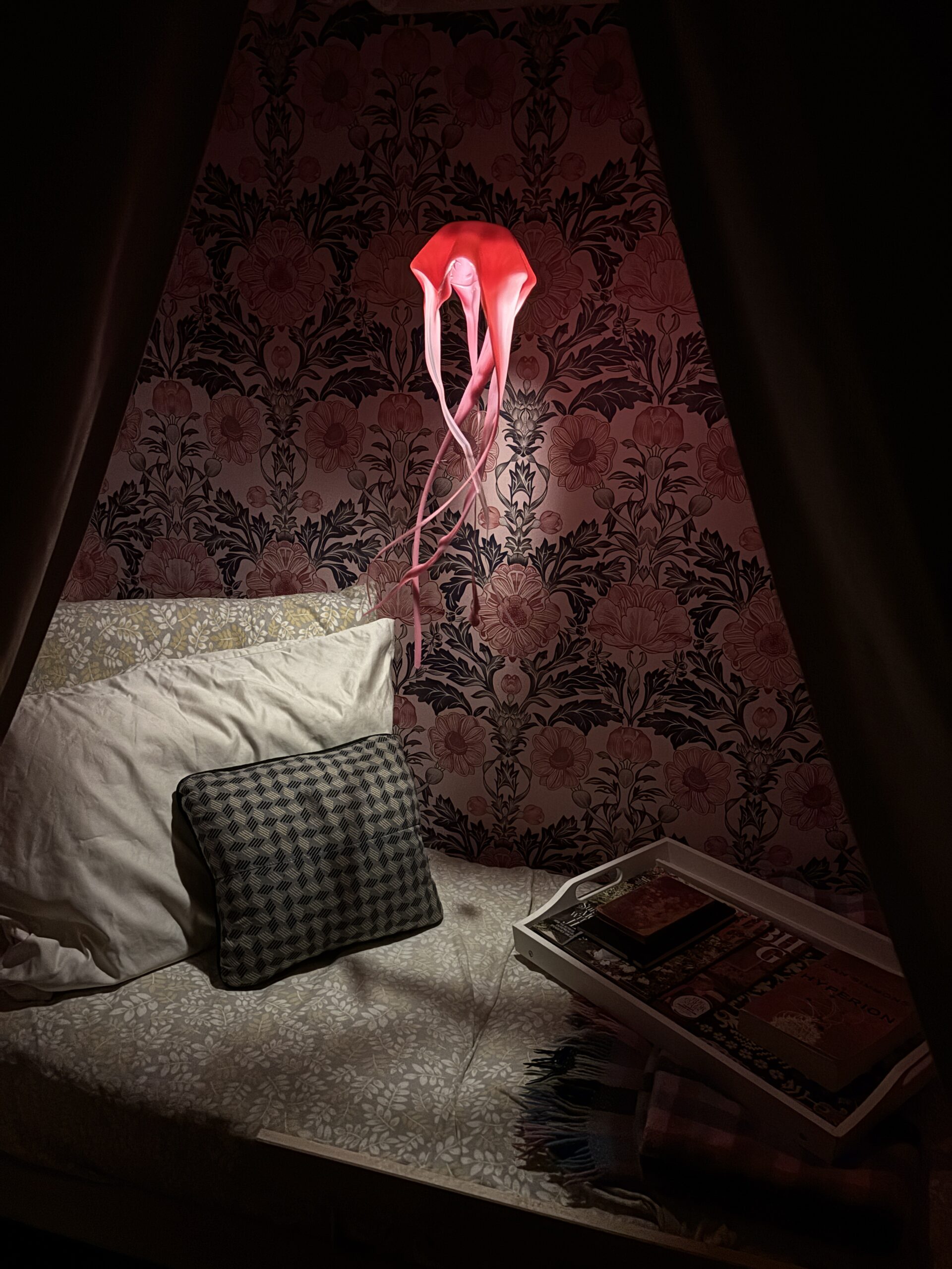 Coral pink jellyfish hanging lamp lighting a cozy bed nook with pillows, books, and floral wallpaper